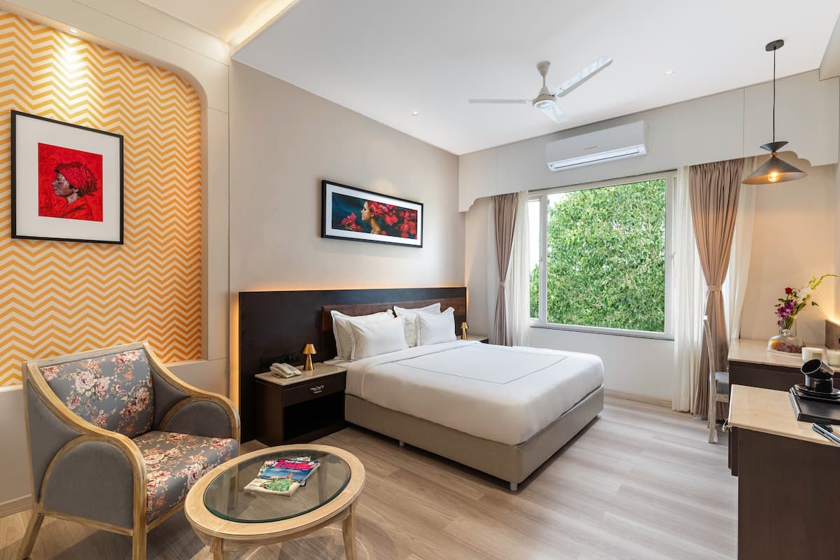 A hotel room with a bed, a chair, a small table, a nightstand, a desk, and a window with curtains.