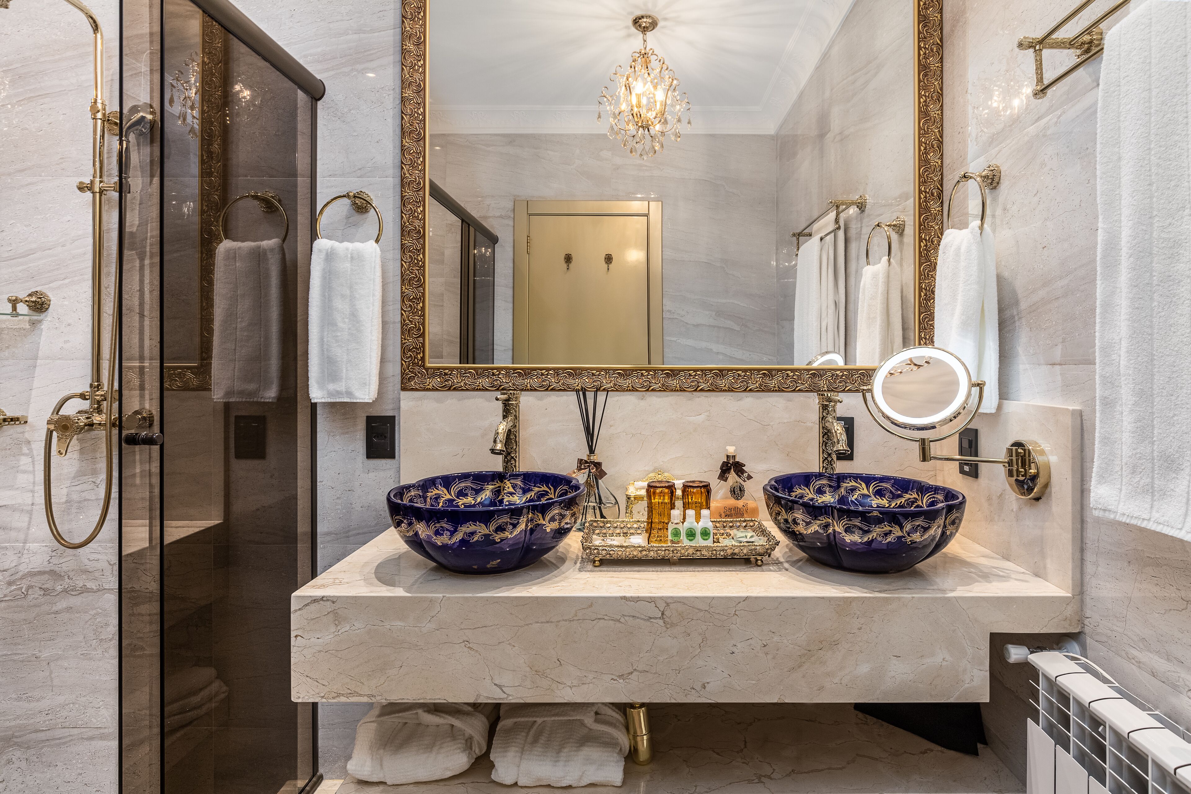 Un baño con lavamanos doble, dos recipientes decorativos grandes, una lámpara de araña y un espejo con marco dorado.