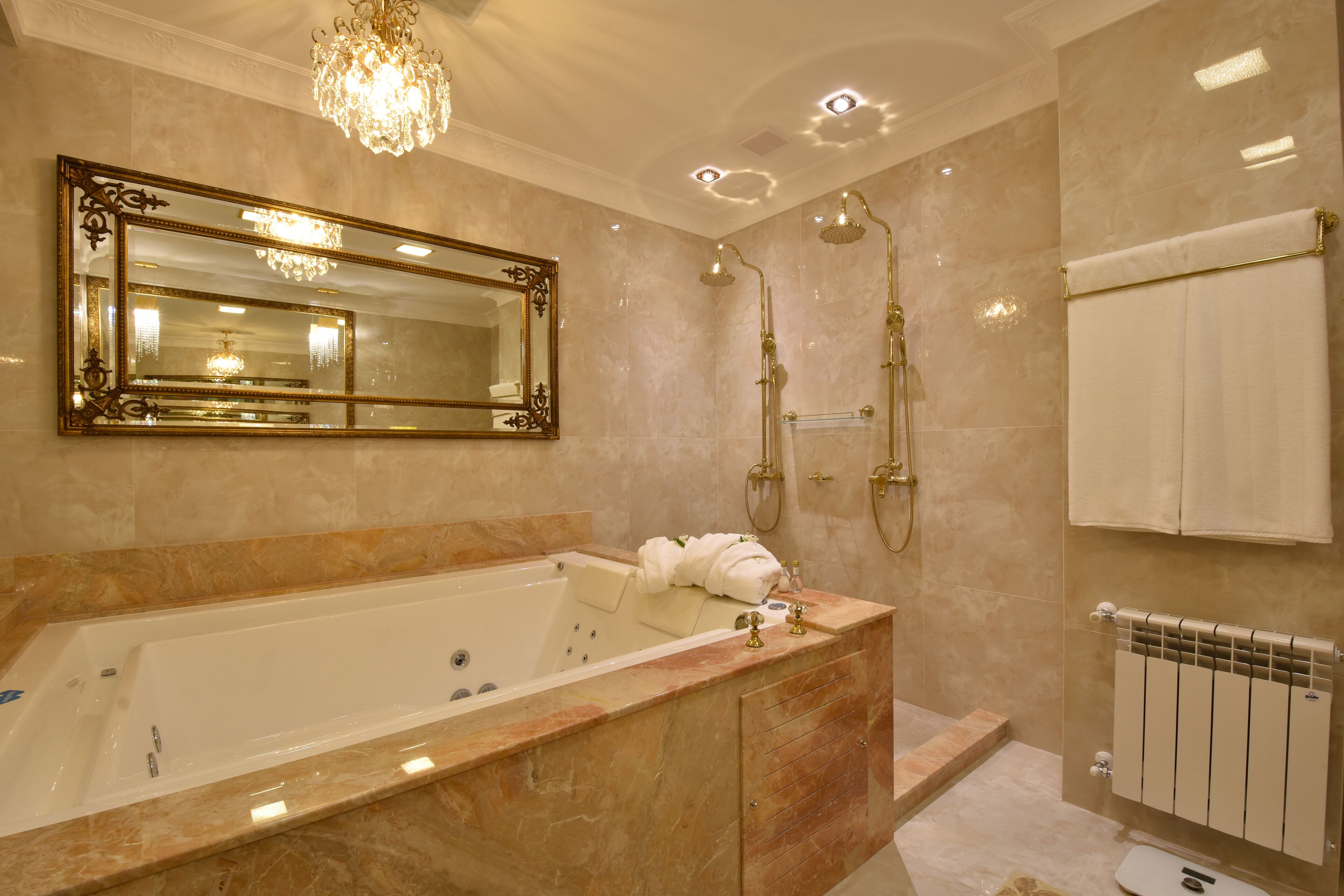 A luxurious bathroom with a large bathtub, a mirror with an ornate frame, a shower with a golden showerhead, and a towel rack with white towels.