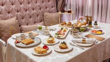 Una mesa dispuesta con diferentes alimentos para el desayuno, como pasteles, sándwiches y bebidas, en una habitación con cabecera capitonada y cortinas.