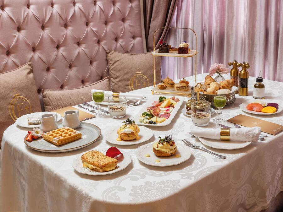 Una mesa dispuesta con diferentes alimentos para el desayuno, como pasteles, sándwiches y bebidas, en una habitación con cabecera capitonada y cortinas.