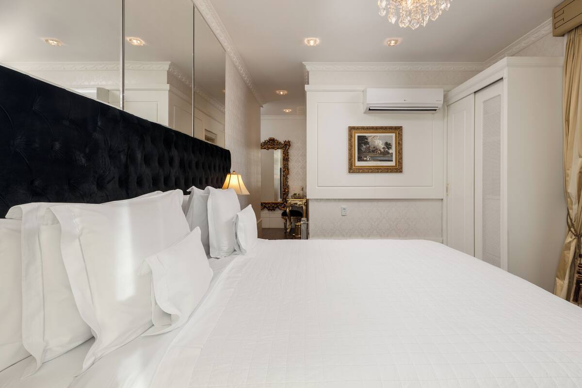 Ett hotellrum med en stor säng, en svart klädd sänggavel, en kristallkrona, en luftkonditioneringsanläggning och en inramad tavla på väggen.