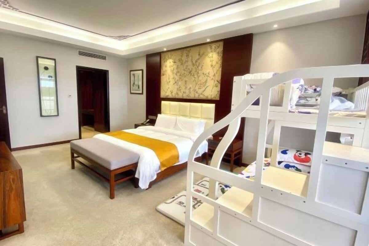 A hotel room with a bed, a bedside table, a headboard, a wall-mounted artwork, a white bunk bed, and a carpeted floor.