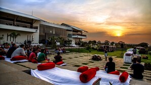 An outdoor seating area with white tables and red cushions, a modern building with a balcony, and a sunset in the background. - Regenta Resort Soma Vine Village (Nashik)