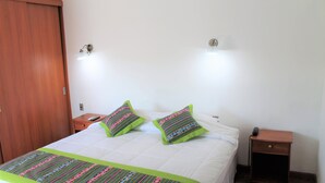 A neatly made bed with a striped bedspread and two pillows, a wooden wardrobe, a bedside table with a phone, and a nightstand with a lamp. - Hotel Samaña (Arica)