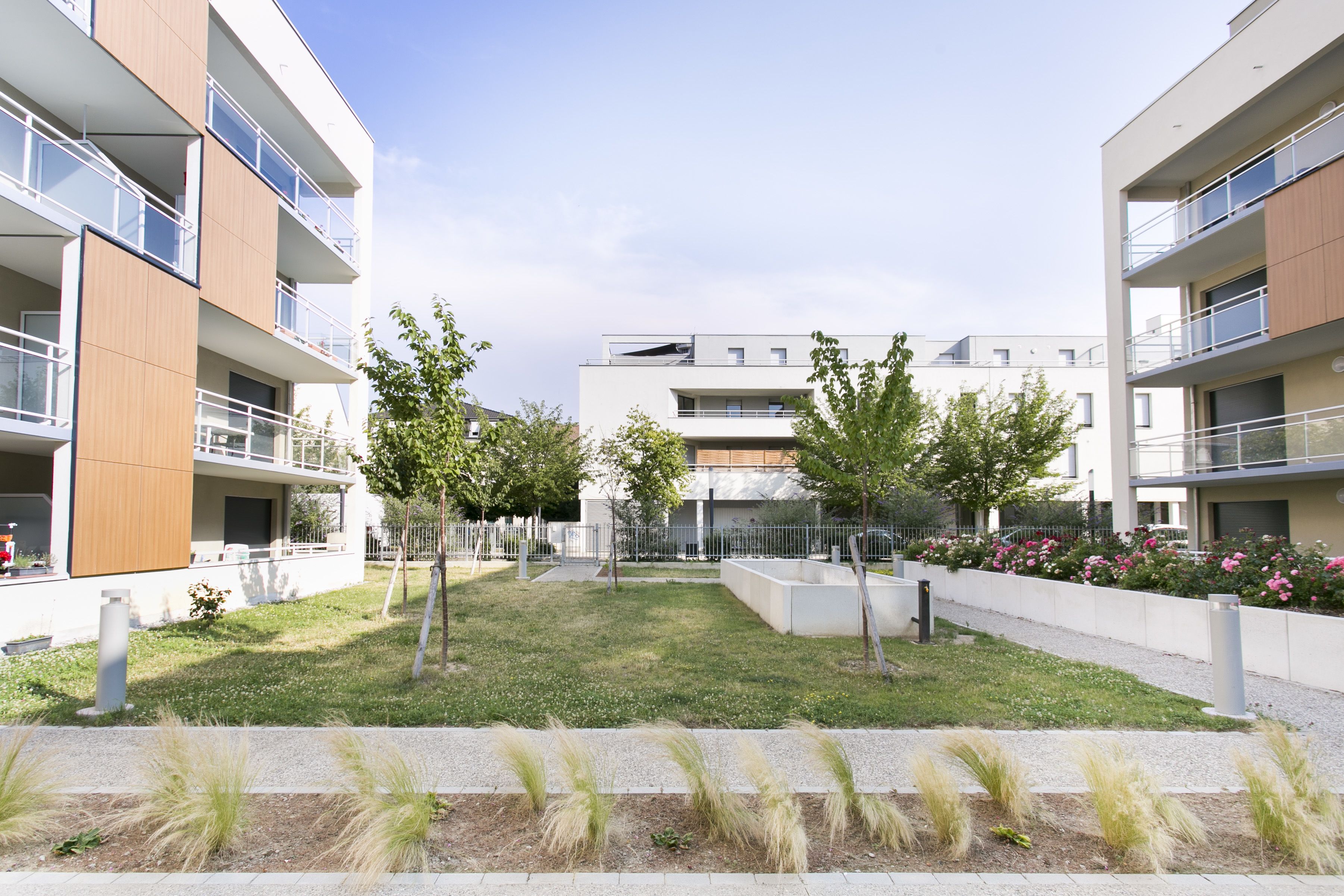 Un moderno complejo de apartamentos con varios balcones, un patio central con césped y árboles, y un macizo de flores de color rosa.