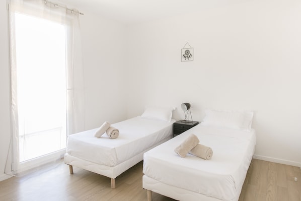 Una habitación con dos camas individuales, una mesita de noche con lámpara y un ventanal con cortinas.