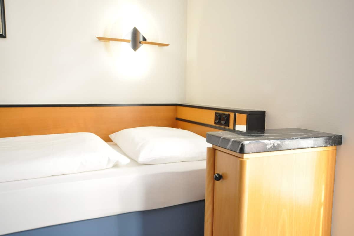 Una habitación de hotel con cama, mesita de noche, lámpara fijada a la pared y un armario pequeño.