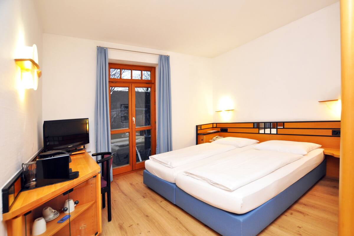 Una habitación de hotel con cama, escritorio con computadora, televisor y ventana con cortinas.