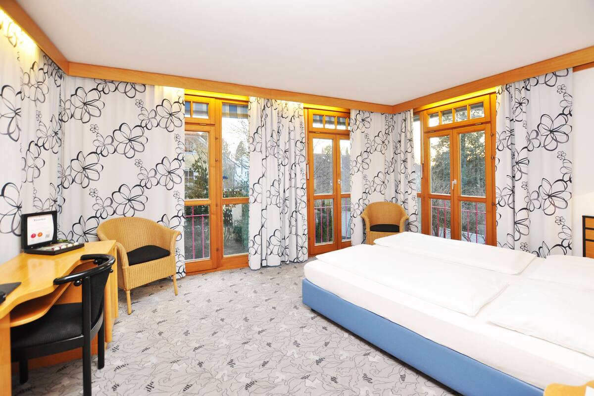 Una habitación de hotel con una cama, un escritorio, dos sillas y ventanas grandes con cortinas.