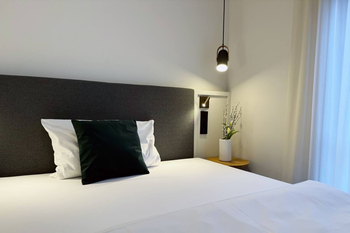 Una habitación de hotel moderna con una cama grande, cabecera oscura, mesita de noche con un jarrón de flores y una lámpara colgante.