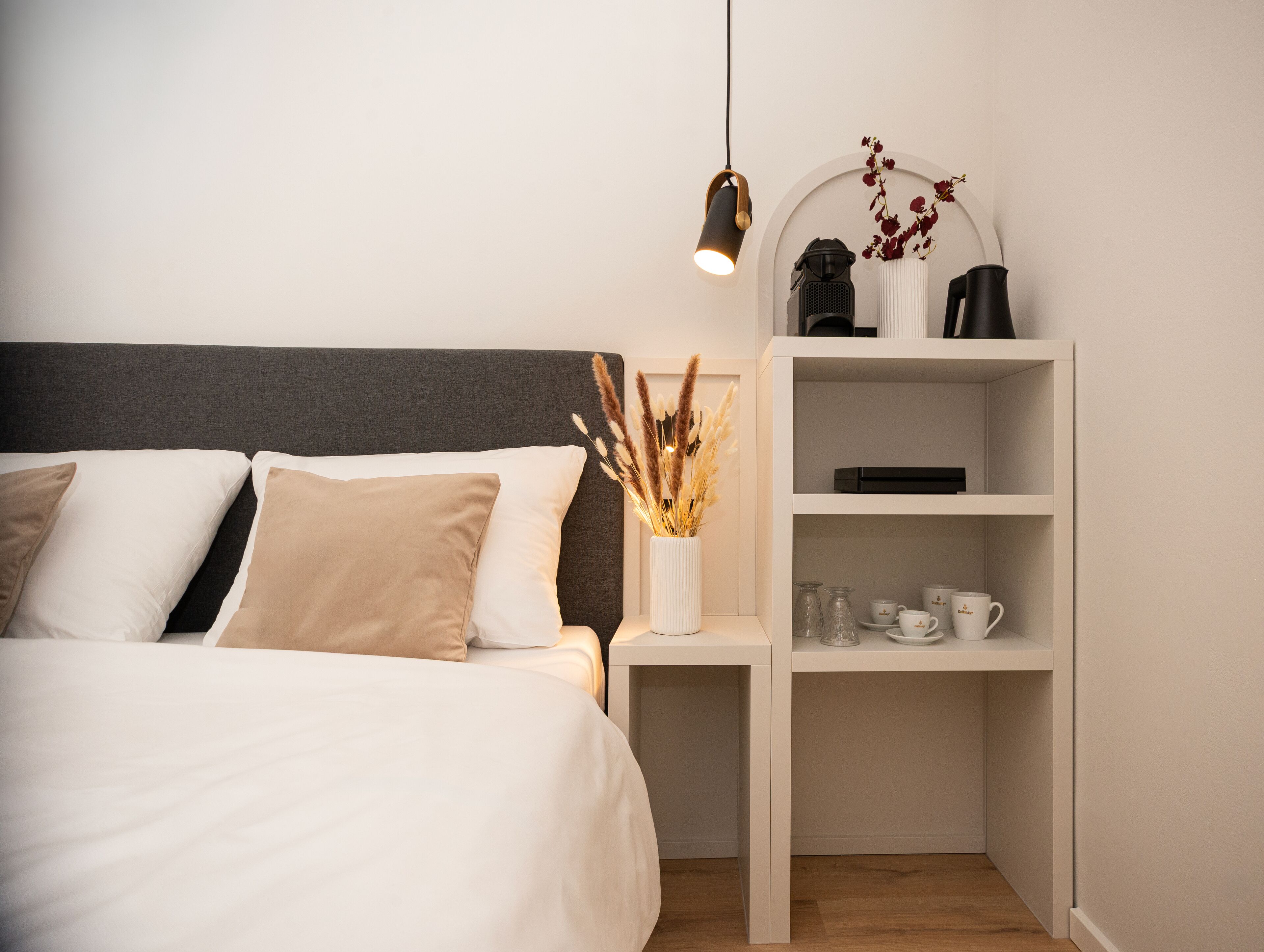A neatly arranged bedroom with a bed, bedside tables, a shelf with cups and a vase, and a hanging light fixture.