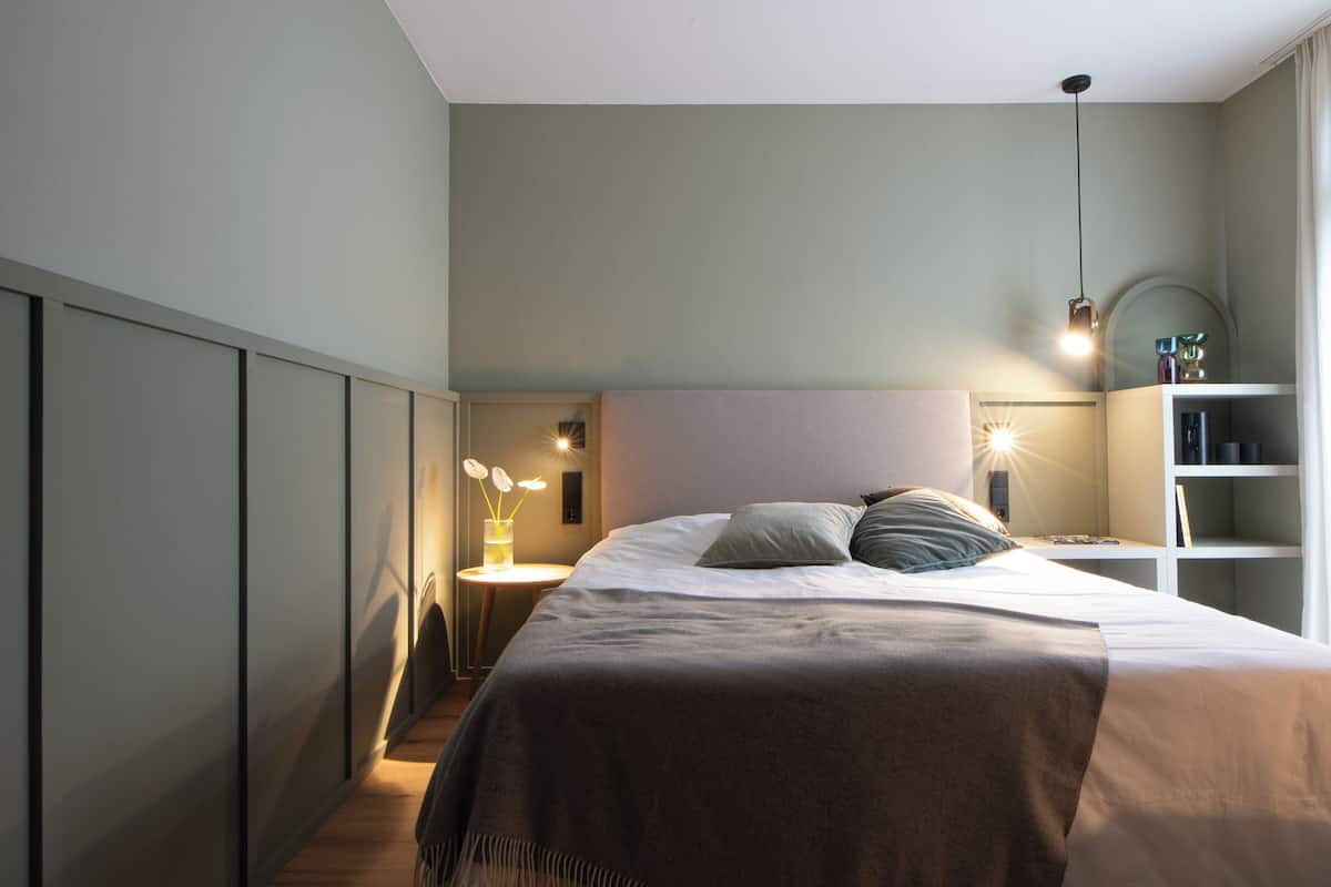 Un dormitorio moderno con una cama grande, una mesita de noche con lámpara, una pequeña mesa redonda con lámpara y una estantería.
