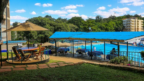 A poolside area with tables and chairs under a blue canopy, surrounded by a pool and greenery. - Methodist Resort (Nairobi)