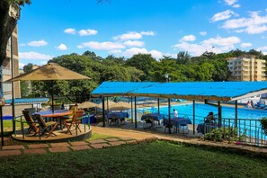 A poolside area with tables and chairs under a blue canopy, surrounded by a pool and greenery.