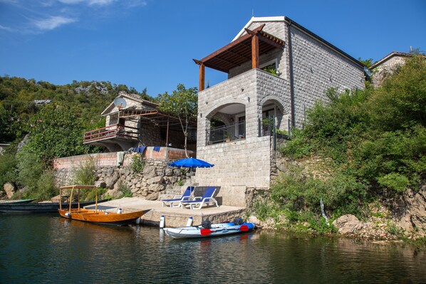 A stone house with a balcony, a dock with boats, and a small waterfall.
