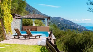 Un área de piscina con sillones de descanso, una pérgola y vistas al mar y a las montañas.