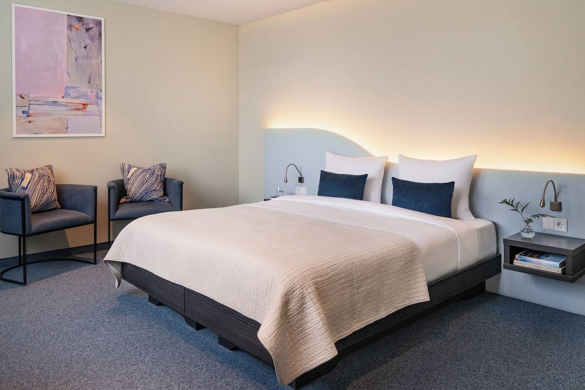 Una habitación de hotel moderna con una cama grande, dos sillones, una mesita de noche y una lámpara de pared.