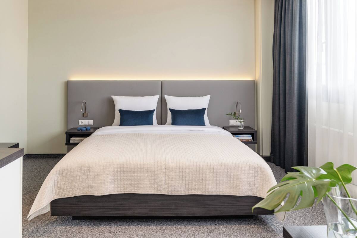 Una cama bien hecha con cabecera, dos mesitas de noche con lámparas y un ventanal con cortinas.
