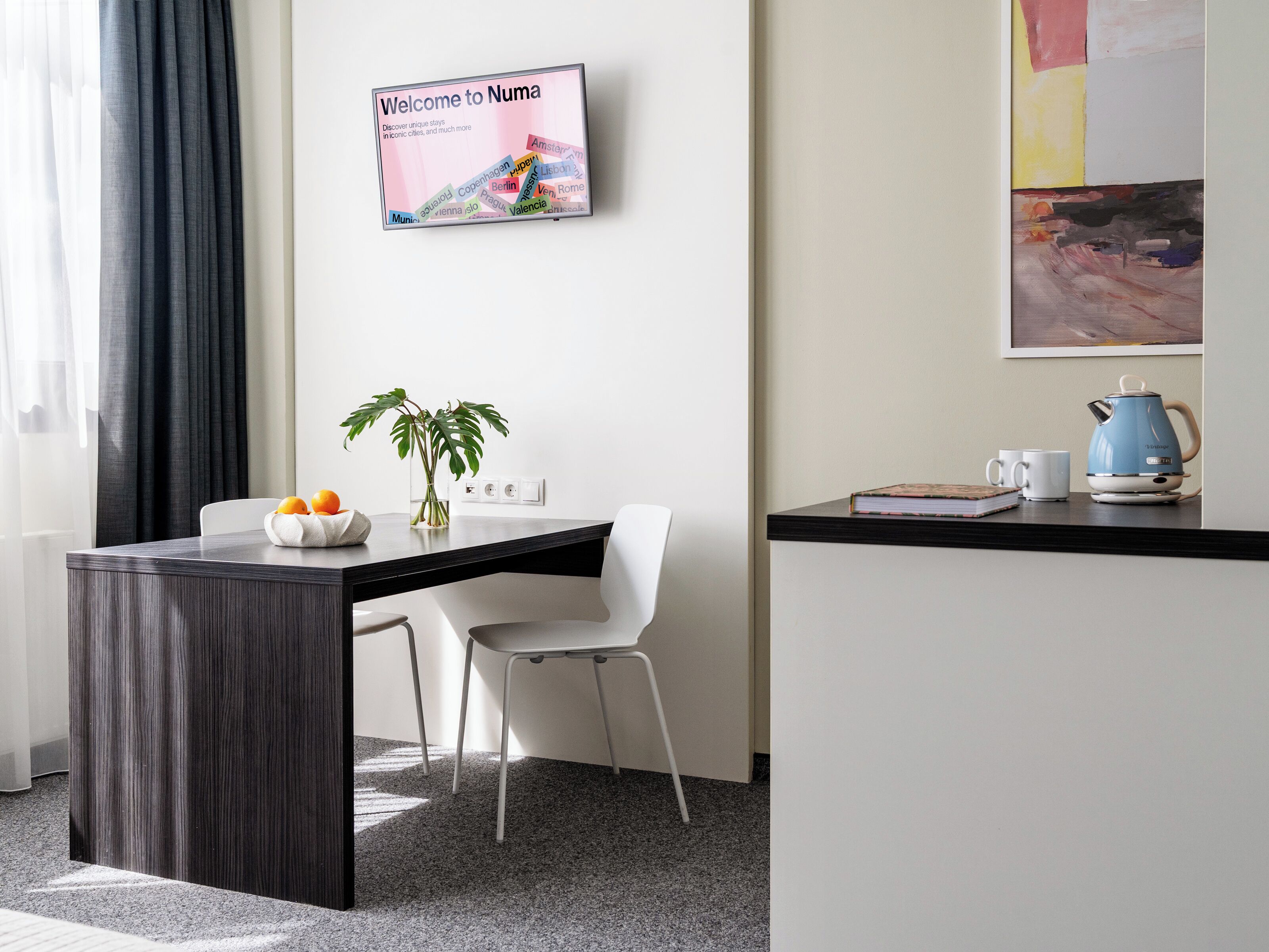 Habitación de hotel moderna con un escritorio de madera oscura, una silla blanca, una planta en maceta, una cafetera y un cartel de bienvenida enmarcado.