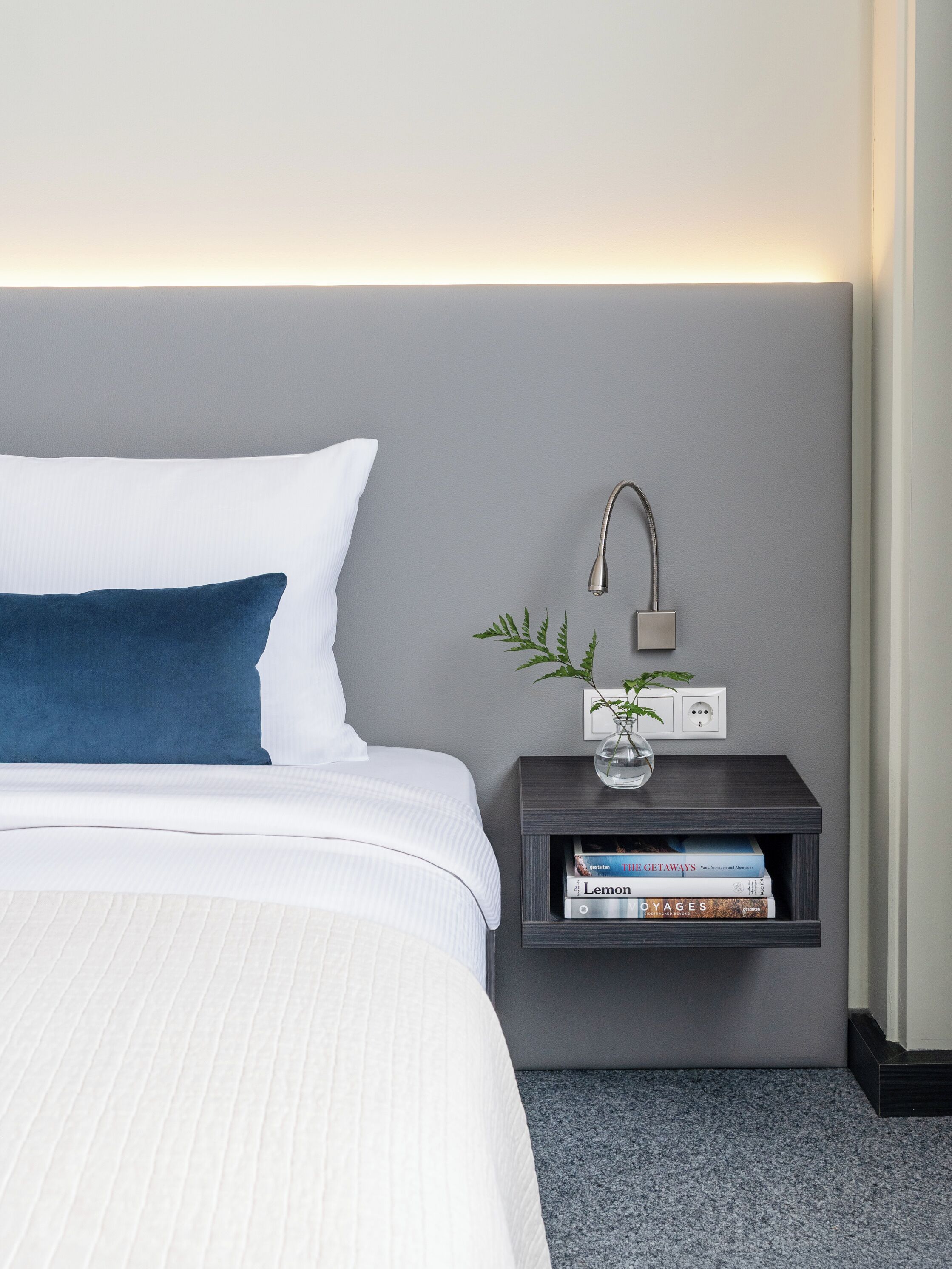 Una cama bien hecha con ropa de cama blanca y azul, una mesita de noche con lámpara y un pequeño estante con libros y una planta.