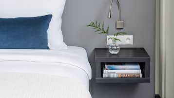 Una cama bien hecha con ropa de cama blanca y azul, una mesita de noche con lámpara y un pequeño estante con libros y una planta.