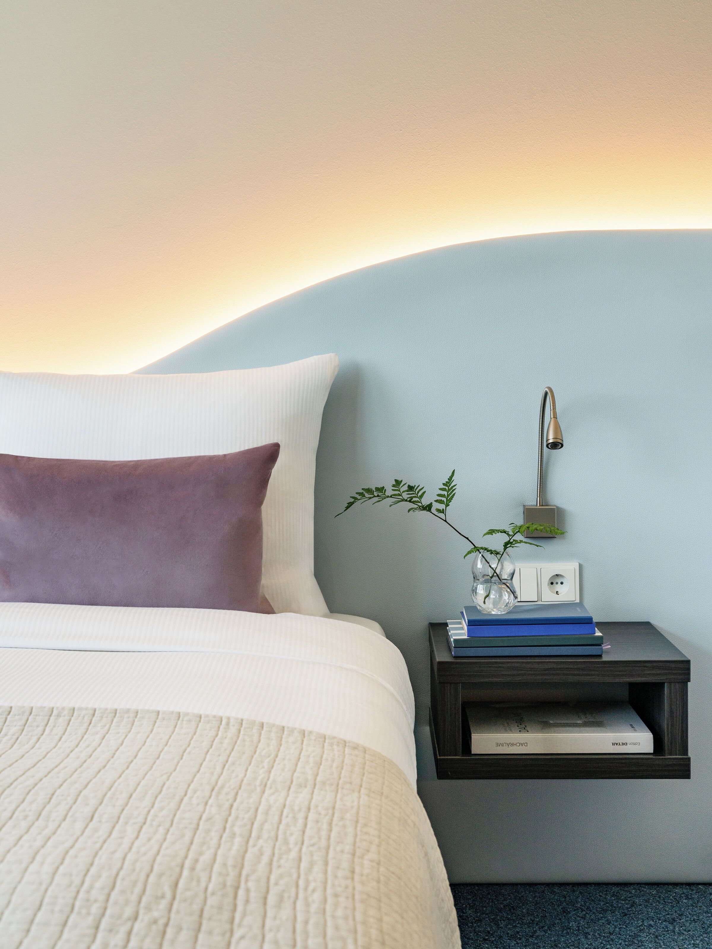 Een keurig opgemaakt bed met een wit en paars kussen, een nachtkastje met een lamp, een kleine plant en een paar boeken.