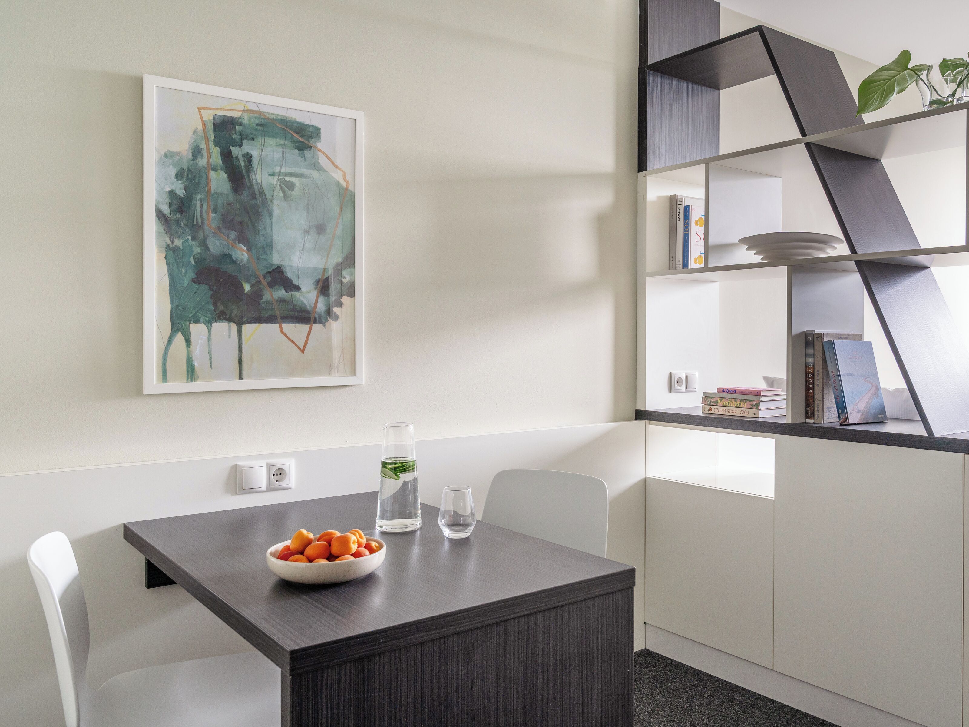 Una cocina moderna con una mesa de madera oscura, sillas blancas, un bol de fruta, un vaso de agua y una estantería con libros y objetos decorativos.