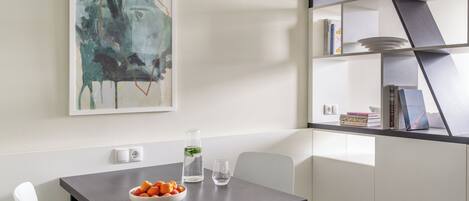 Una cocina moderna con una mesa de madera oscura, sillas blancas, un bol de fruta, un vaso de agua y una estantería con libros y objetos decorativos.
