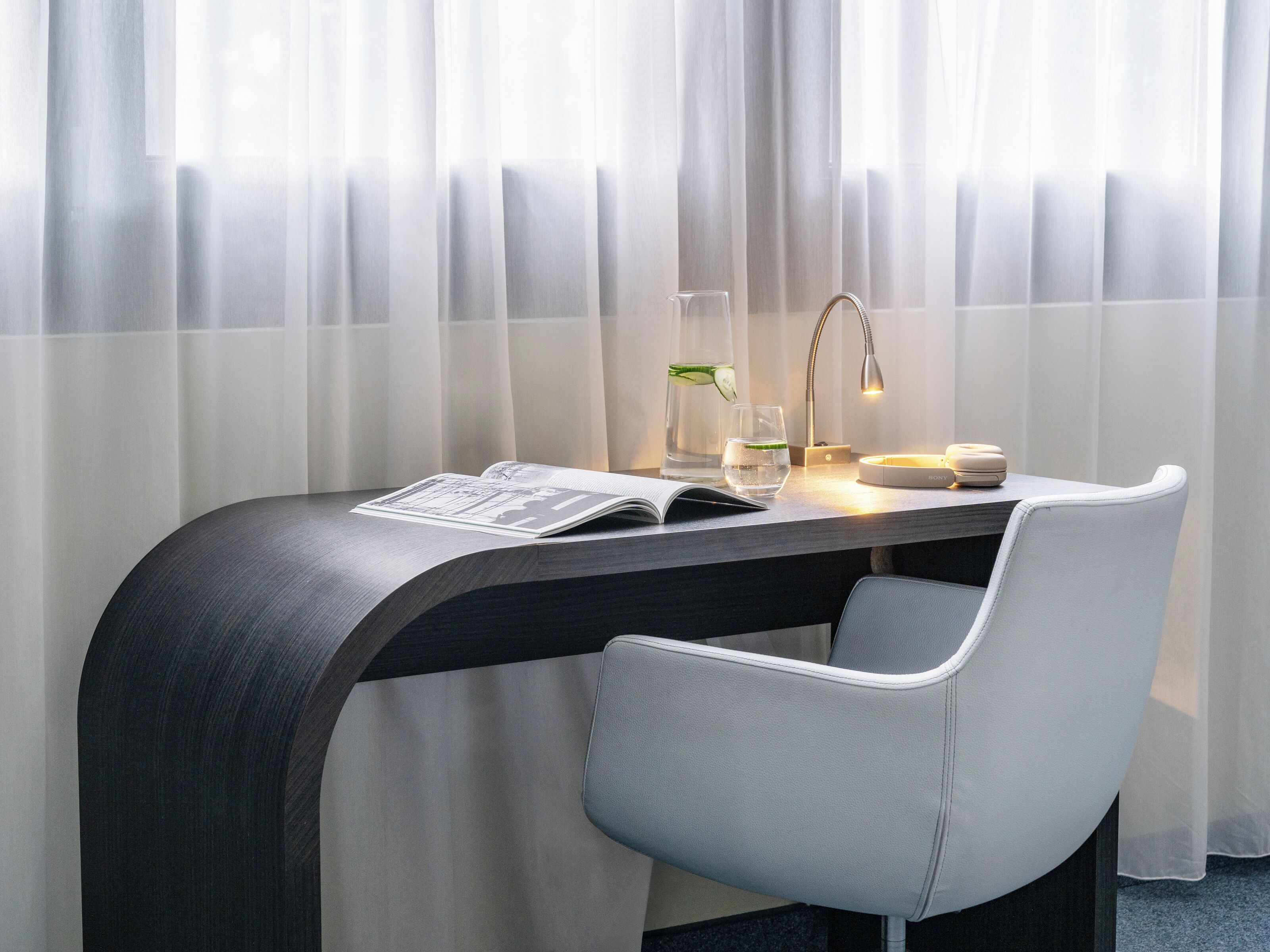A modern hotel room with a curved desk, a chair, a lamp, a glass of water, and a book.