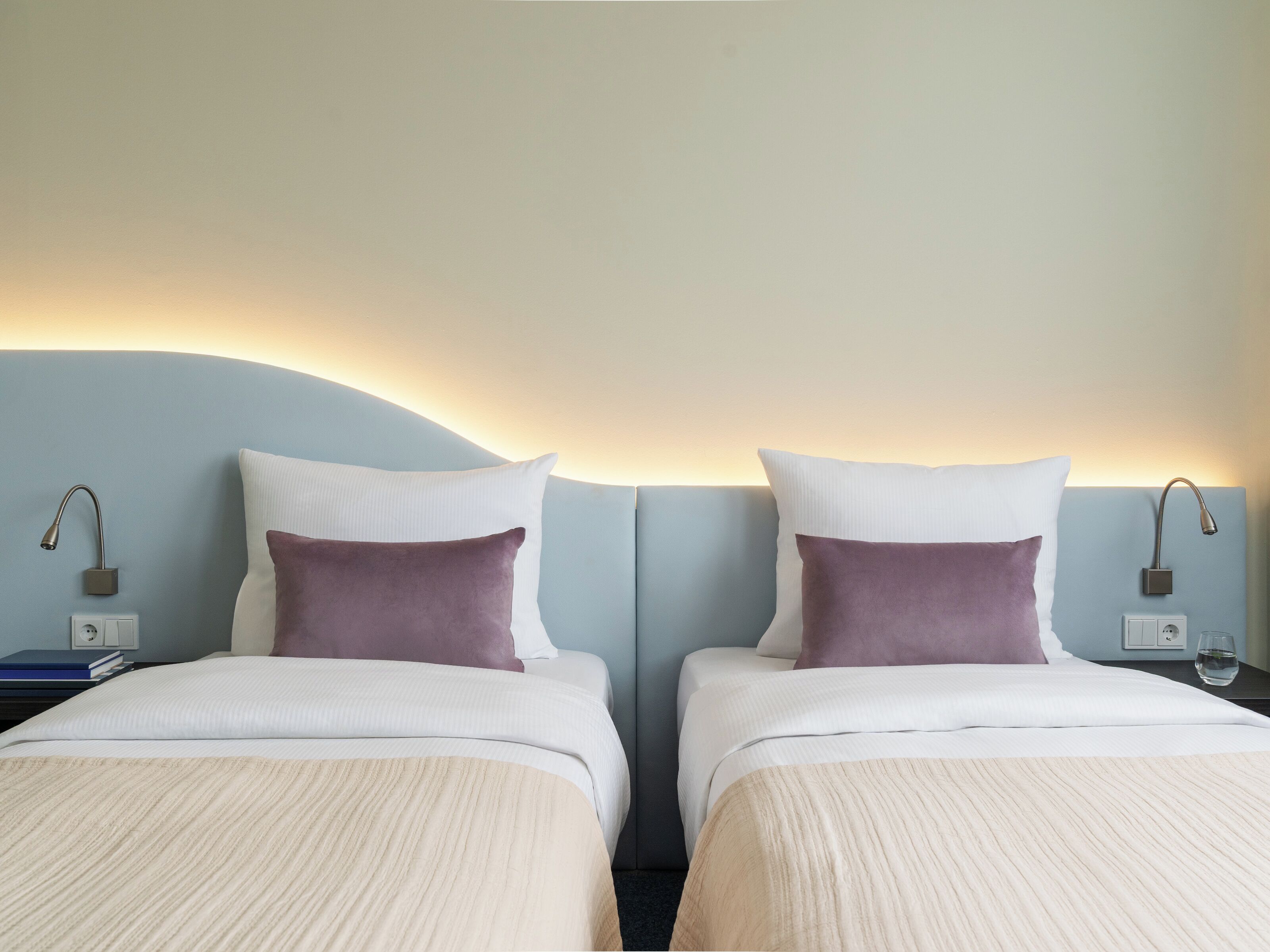Une chambre d’hôtel avec deux lits, chacun agrémenté d’un oreiller blanc et violet, et une table de chevet avec une lampe.