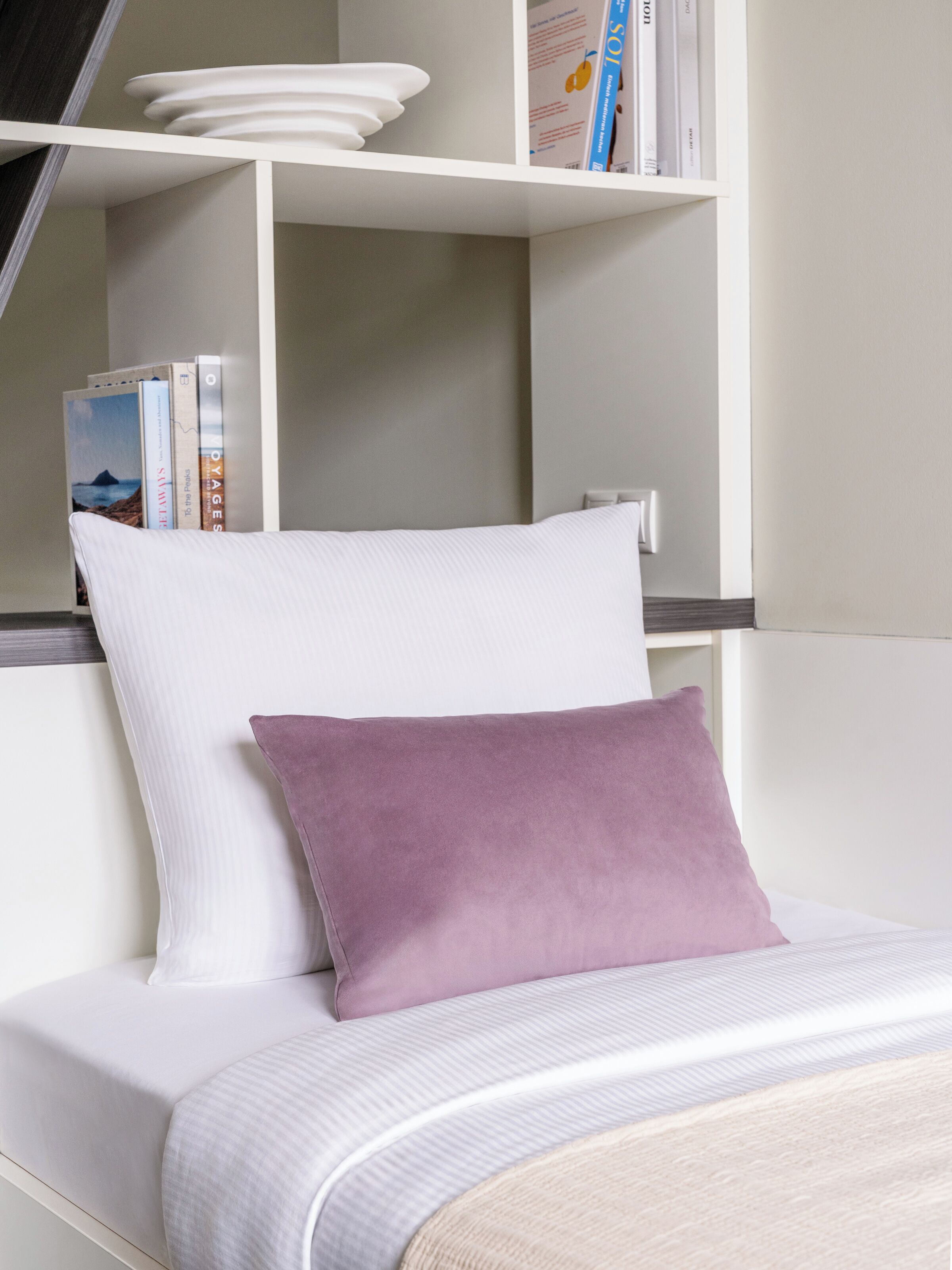 A neatly made bed with a white mattress and a single purple pillow, a white shelf with books and a plate, and a plain wall.