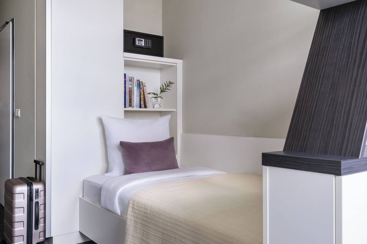 Habitación de hotel pequeña con una cama individual, una mesita de noche, una estantería con libros y una planta, una maleta y una puerta con manija.