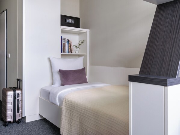 Une chambre d’hôtel de taille réduite, avec un lit simple, une petite table de chevet, une bibliothèque avec des livres et une plante, une valise et une porte avec une poignée.