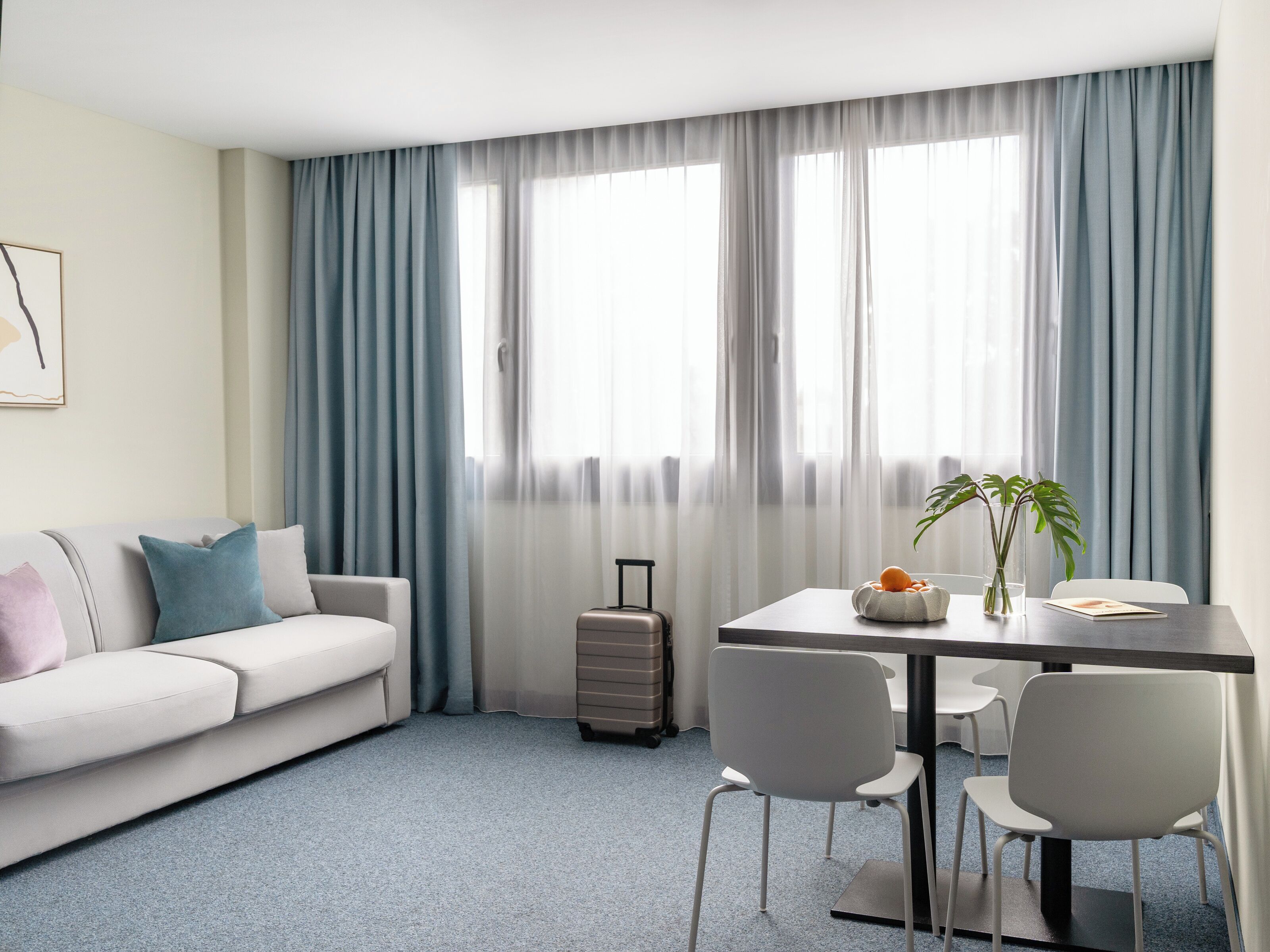 A modern hotel room with a sofa, a dining table with chairs, a suitcase, and a plant.