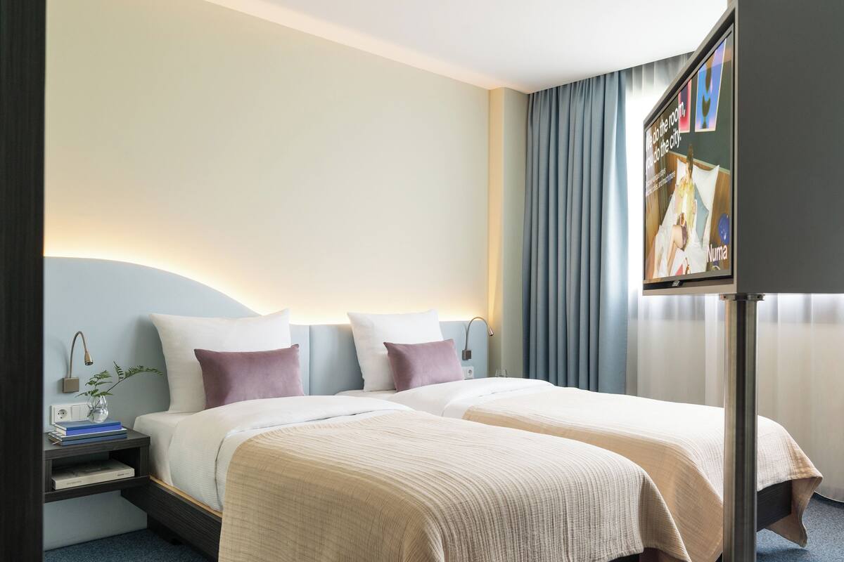 Habitación de hotel con dos camas, una mesita de noche con lámpara, una pequeña mesa con un jarrón y libros, y un ventanal con cortinas.