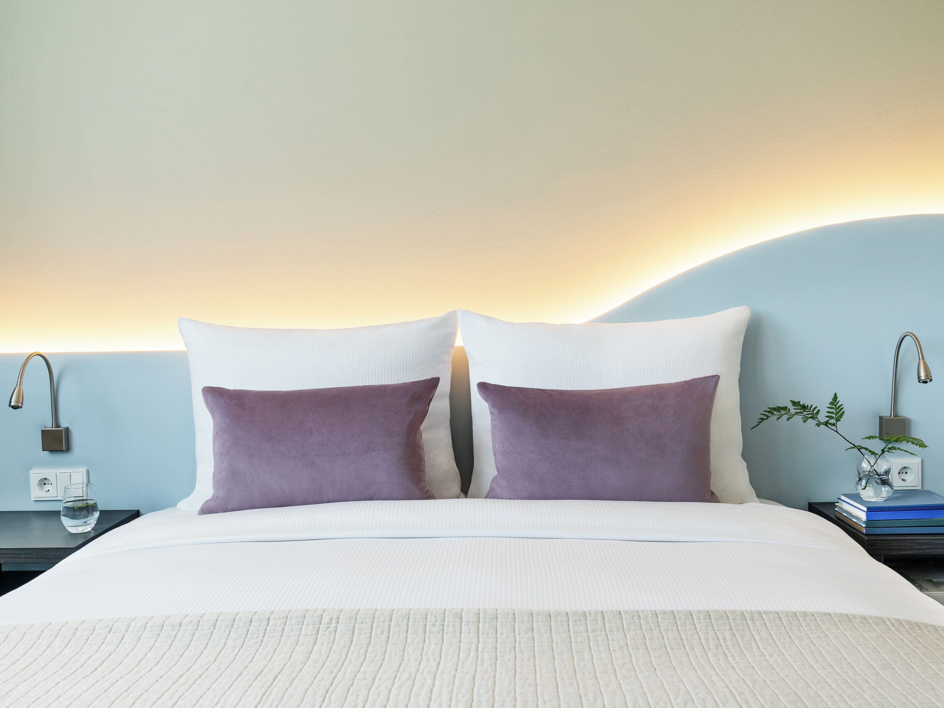 Et hotelværelse med en seng, to puder, et natbord med en lampe, en lille plante og et sengebord med en bog og et glas.