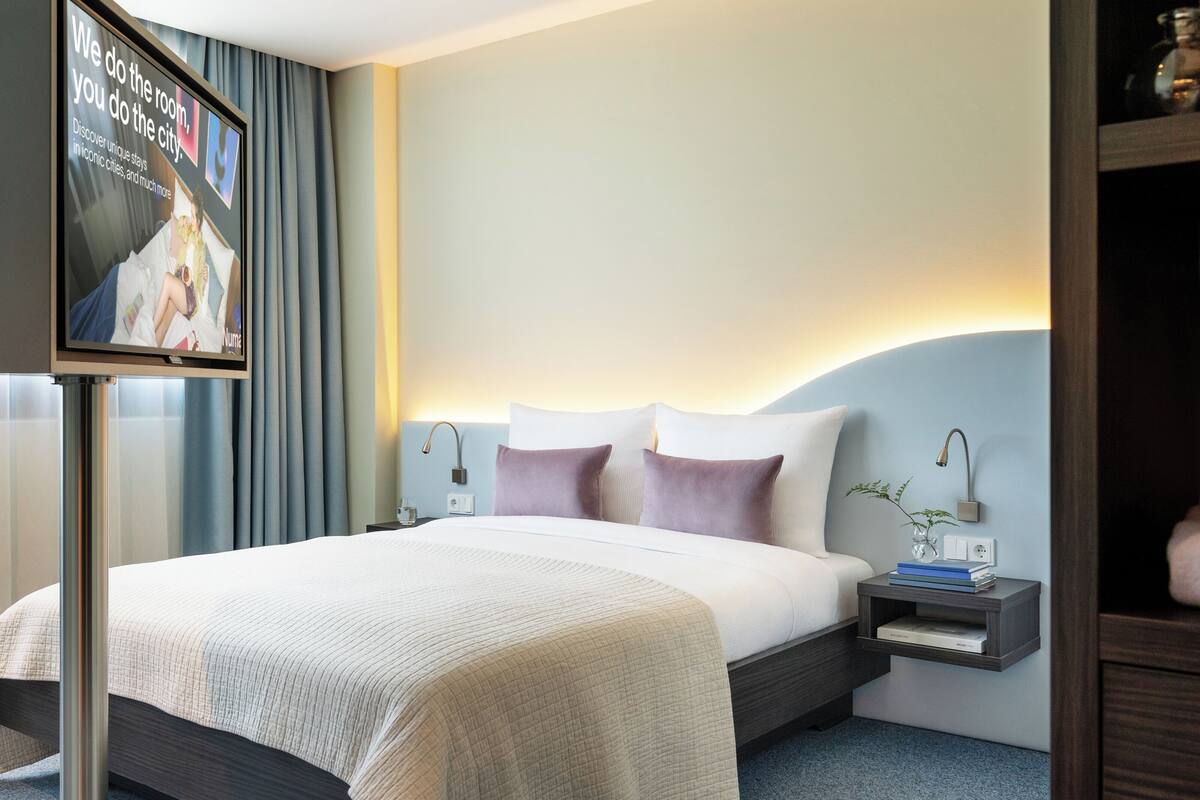 Una habitación de hotel moderna con una cama grande, una mesita de noche con una lámpara, una pequeña mesita de noche con un jarrón y un televisor empotrado en la pared que muestra una imagen promocional.