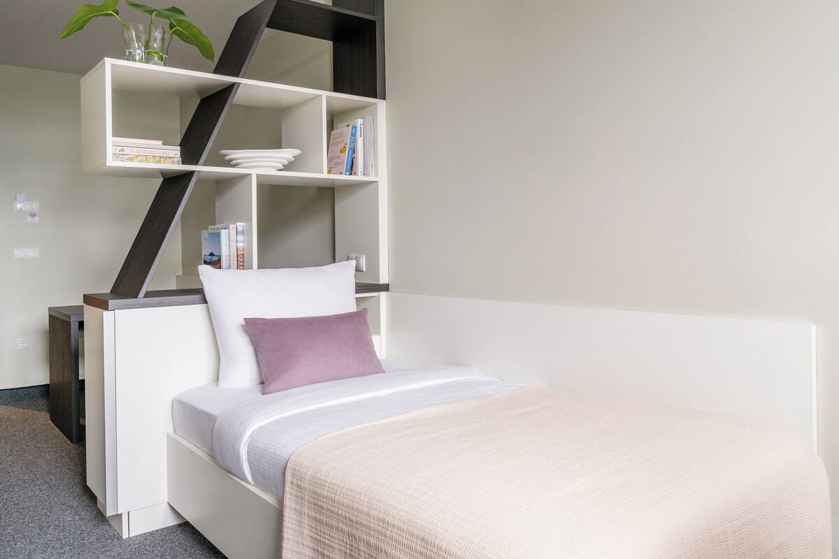 Un dormitorio moderno con una cama blanca, un cojín morado, una manta beige y una estantería empotrada con una planta.