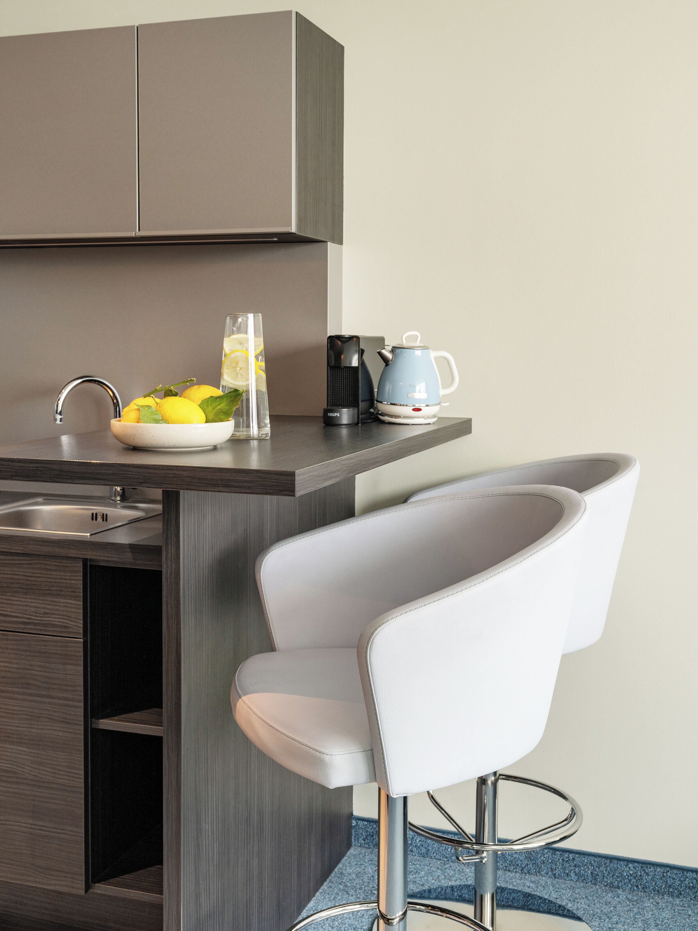 Une cuisine moderne avec un évier encastré, un bol de citrons, un verre d’eau, une machine à café et une chaise.