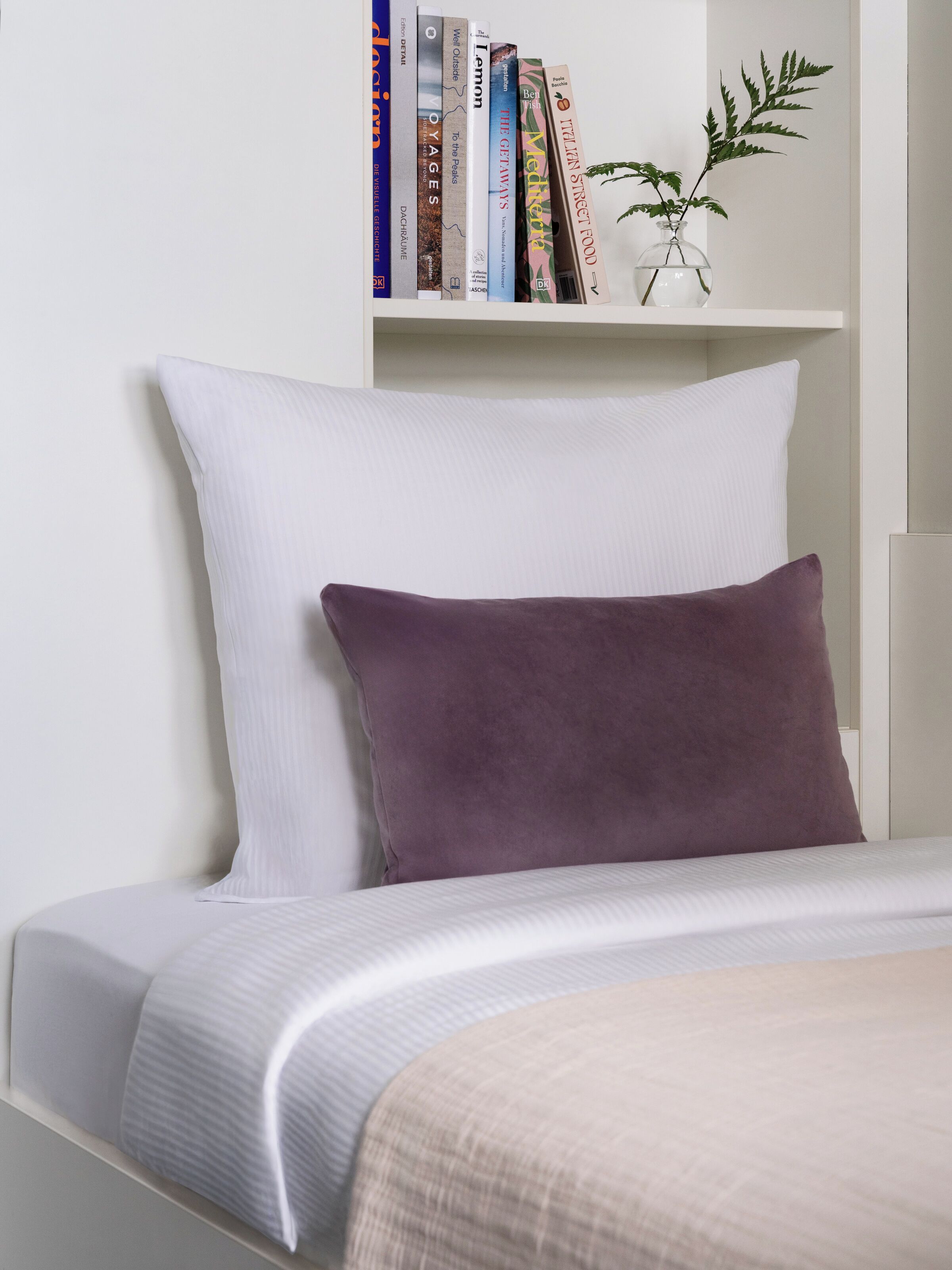 A neatly made bed with a white and purple pillow, a white headboard, and a white bedside table.
