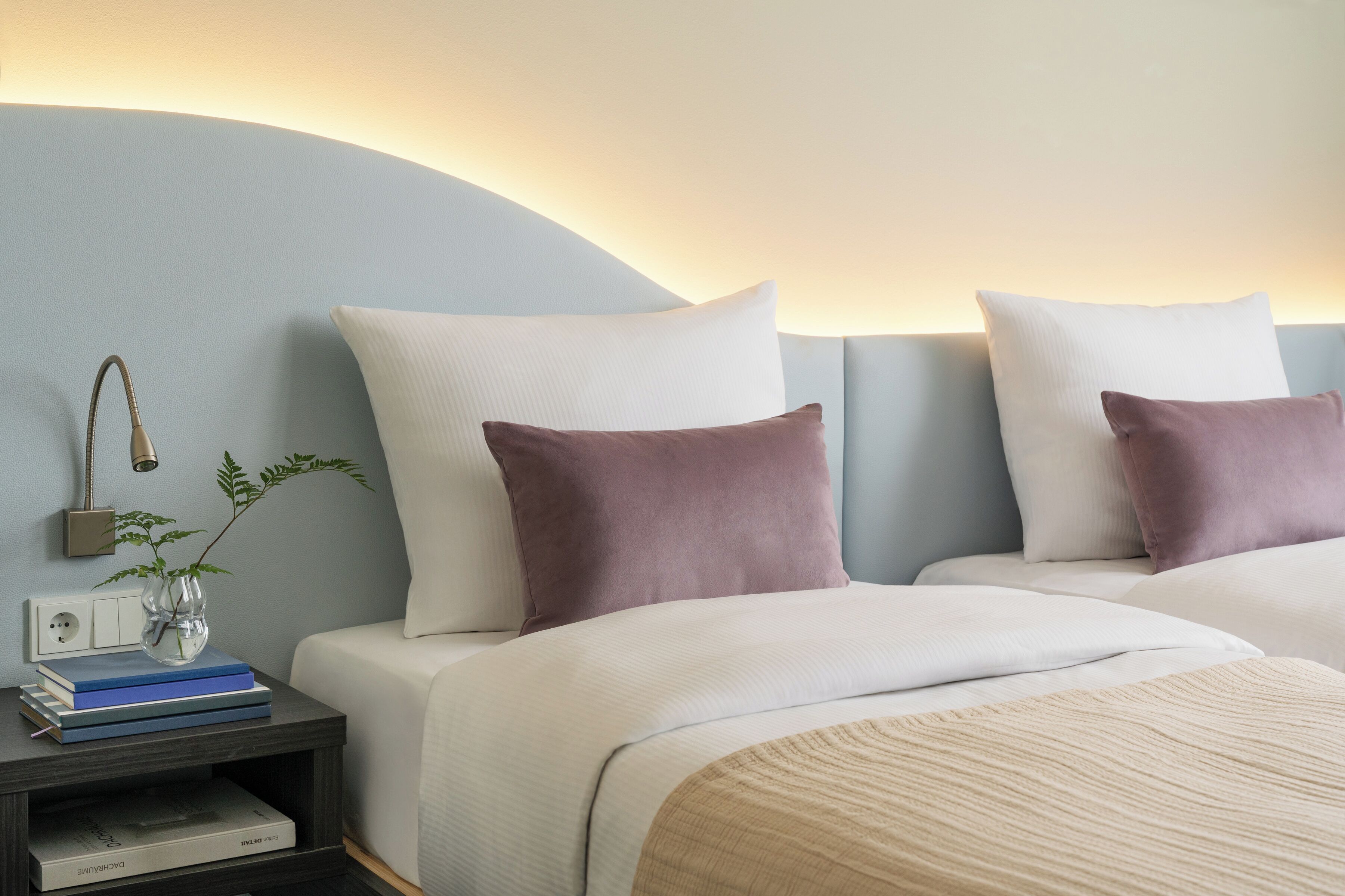 A hotel room with two beds, a nightstand with books and a vase, and a wall-mounted lamp.
