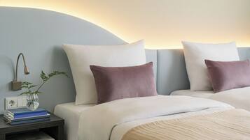 A hotel room with two beds, a nightstand with books and a vase, and a wall-mounted lamp.
