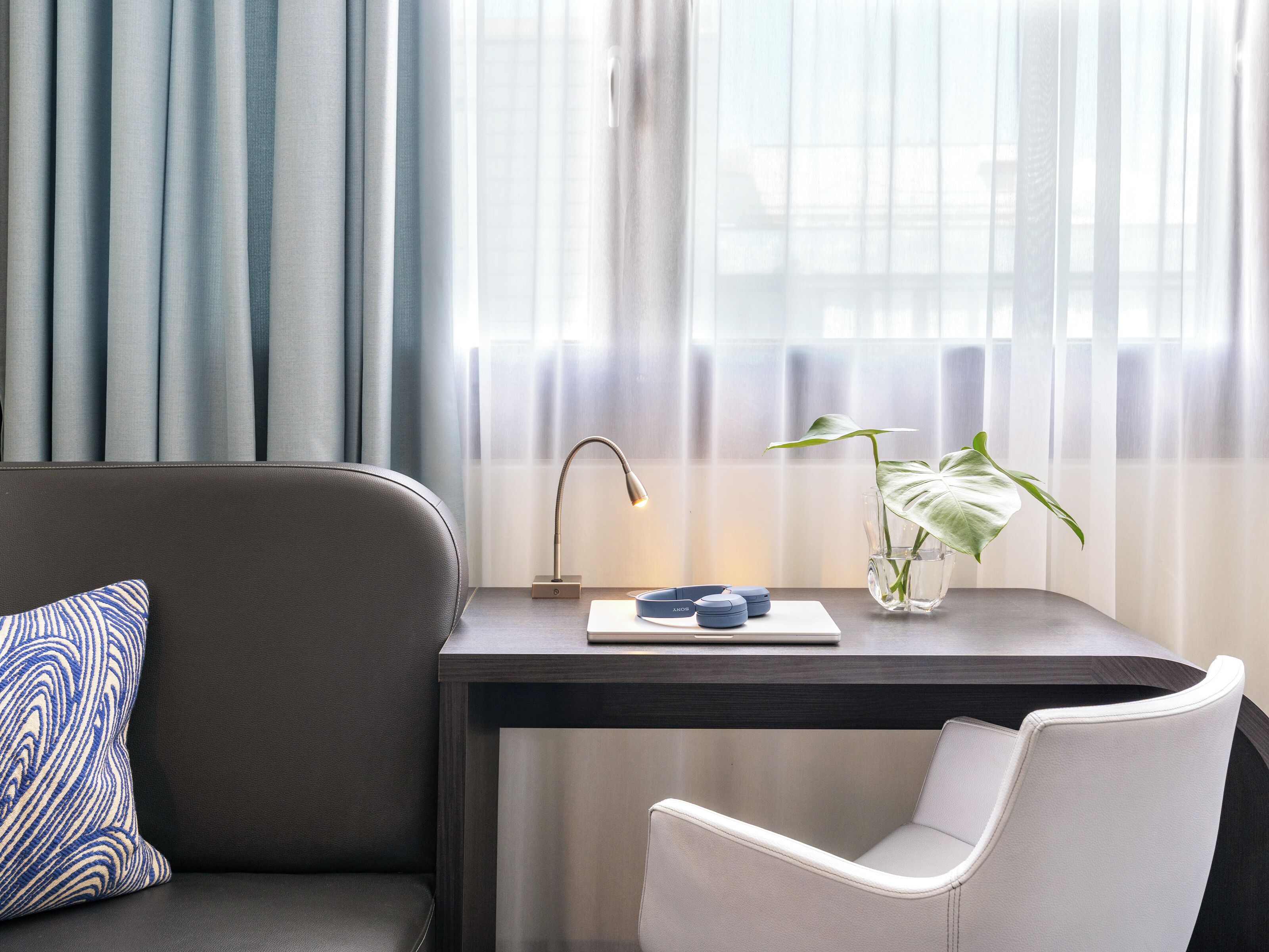 A modern room with a grey sofa, a white chair, a desk with a lamp, a plant, and a window with sheer curtains.