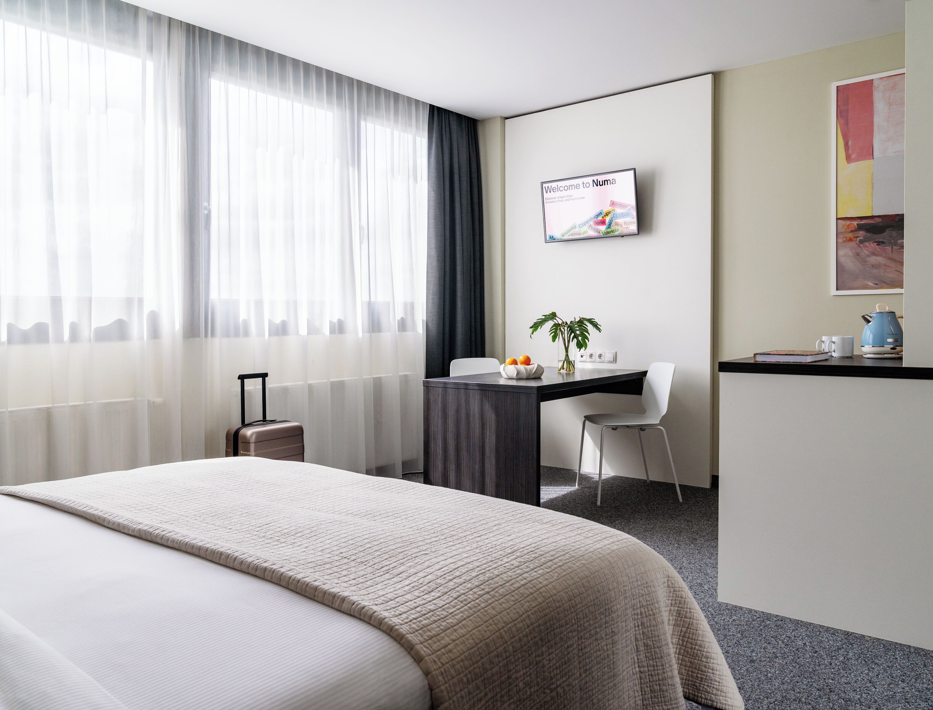 Habitación de hotel con cama, escritorio, silla, una mesita con una planta, tetera y un cartel de bienvenida enmarcado.