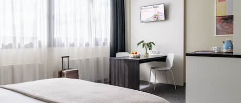 Habitación de hotel con cama, escritorio, silla, una mesita con una planta, tetera y un cartel de bienvenida enmarcado.