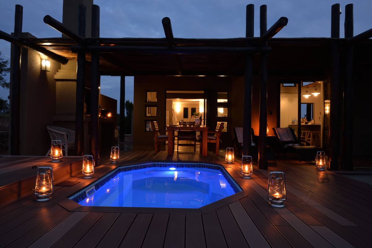 Un bain à remous entouré d’une terrasse en bois et éclairé par des lanternes, avec un patio couvert et une vue sur l’intérieur de la maison.