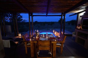 An outdoor dining area with a wooden table set for two, surrounded by chairs, and a pool in the background.