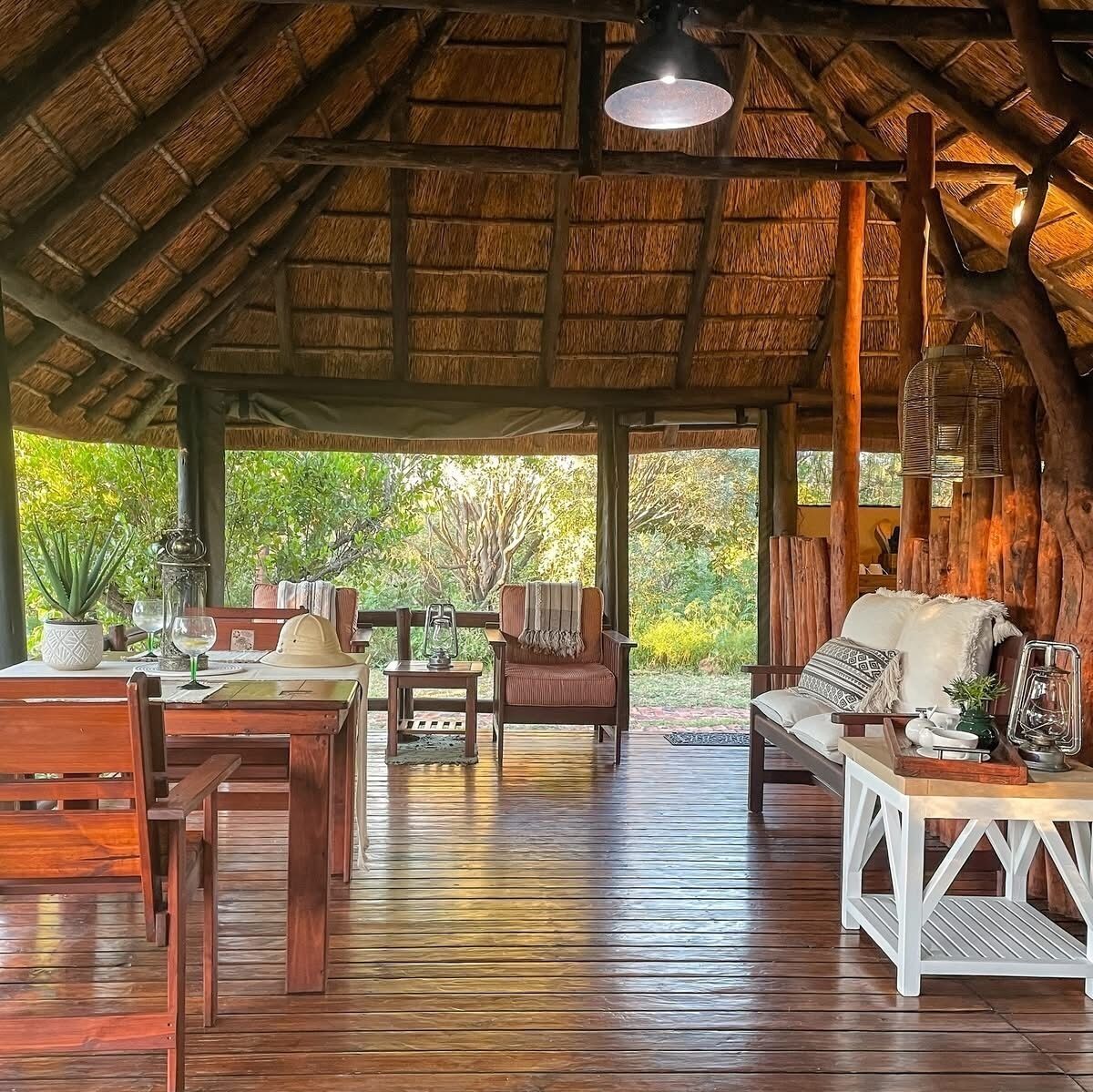 An open-air lounge with wooden furniture, a thatched roof, and a view of greenery outside.