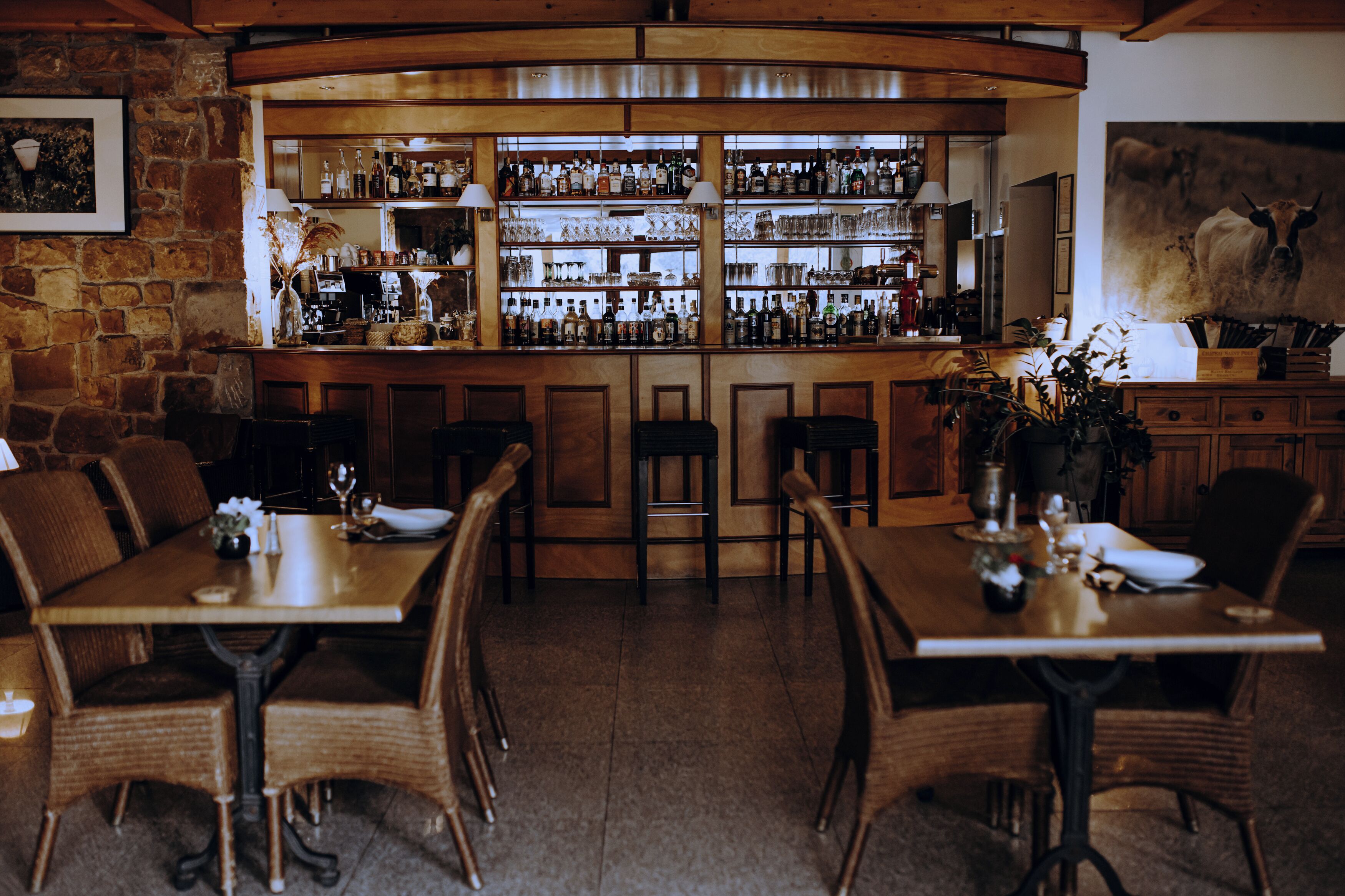 Een bar met flessen op planken, een groot schilderij van een koe en houten meubels.
